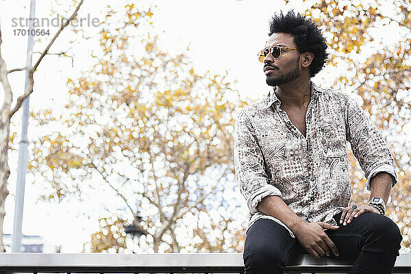Stilvoller erwachsener Mann mit Afro-Haar und Sonnenbrille auf dem Geländer sitzend