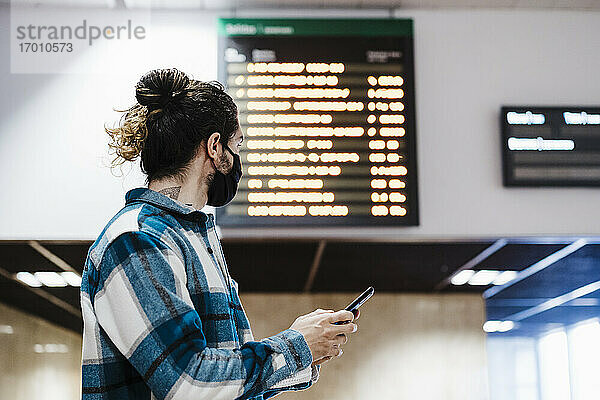 Junger Mann mit Gesichtsmaske  der ein Mobiltelefon benutzt  während er in einer U-Bahn-Station wartet