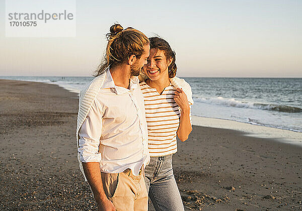 Lächelnde junge Frau  die mit ihrem Freund am Strand bei Sonnenuntergang spazieren geht