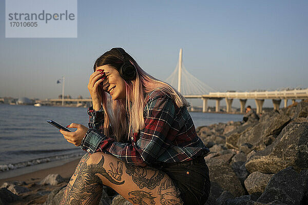 Lächelnde junge Frau mit Smartphone hört Musik auf Felsen am Strand an einem sonnigen Tag