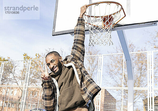 Glücklicher junger Mann  der Frieden gestikuliert  während er an einem sonnigen Tag am Basketballkorb hängt