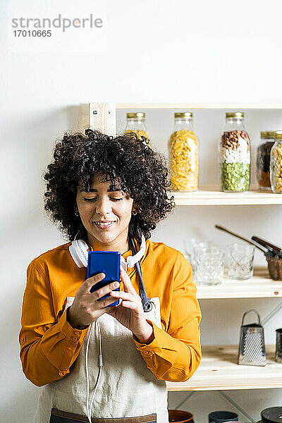 Frau mit lockigem Haar  die ein Mobiltelefon benutzt  während sie in der Küche zu Hause steht