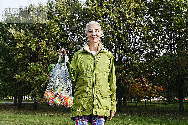Ältere Frau hält eine mit Obst und Gemüse gefüllte Netztasche in einem öffentlichen Park