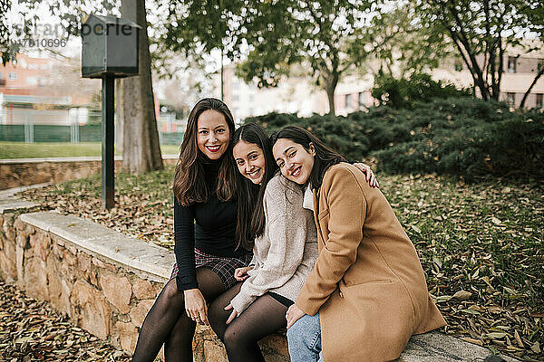 Lächelnde reife Frau mit Töchtern  die auf einer Stützmauer im Park sitzen