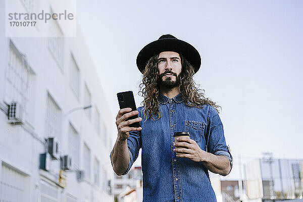 Stilvoller Mann mit Einweg-Kaffeebecher schaut weg  während er sein Smartphone in der Stadt hält