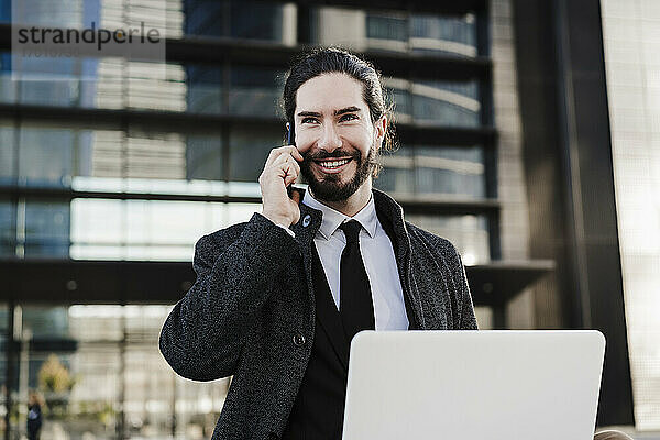 Lächelnder Geschäftsmann mit Laptop  der im Freien sitzend mit seinem Mobiltelefon spricht