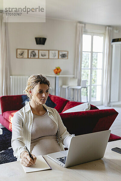 Freiberuflerin  die in ein Buch schreibt  während sie einen Laptop am Schreibtisch im Wohnzimmer benutzt