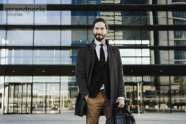 Männlicher Unternehmer mit Händen in der Tasche  der eine Aktentasche trägt  während er vor einem modernen Gebäude steht