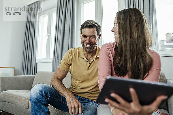 Reife Frau mit digitalem Tablet  die einen glücklichen Mann ansieht  während sie auf dem Sofa in einer Wohnung sitzt