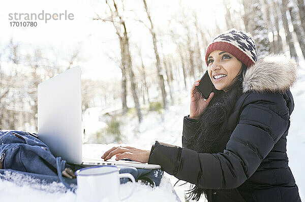 Lächelnde Geschäftsfrau mit Laptop  die mit ihrem Smartphone spricht  während sie auf einer Bank im Schnee sitzt