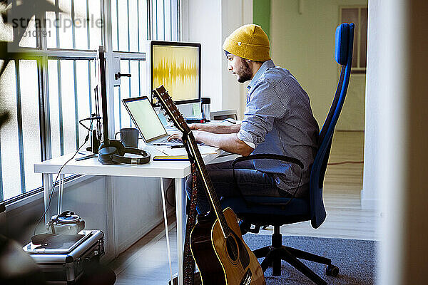 Musiker arbeitet am Laptop  während er im Studio an der Gitarre sitzt