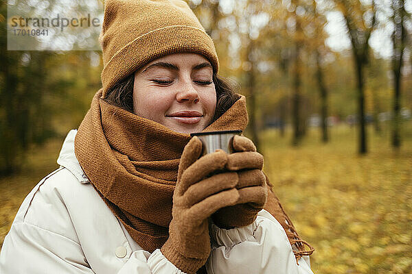 Lächelnde junge Frau mit geschlossenen Augen riecht Tee im Herbst Park