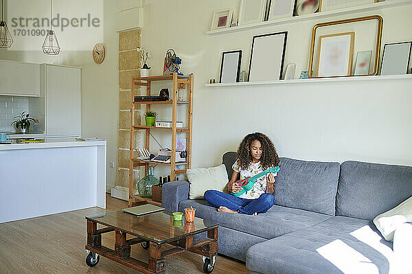 Junge Afro-Frau spielt Ukulele  während sie zu Hause auf dem Sofa sitzt
