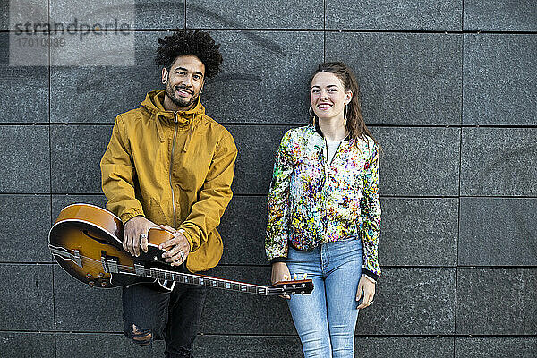 Lächelnder Mann  der eine Gitarre hält  während er neben einer Frau an einer Betonwand steht