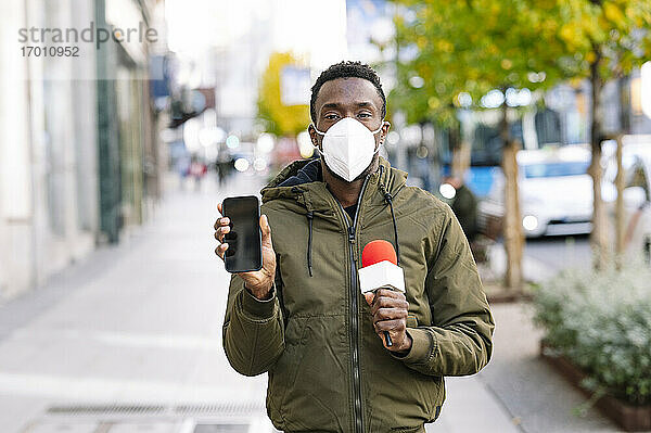 Männlicher Reporter mit Gesichtsmaske zeigt sein Smartphone  während er auf der Straße steht