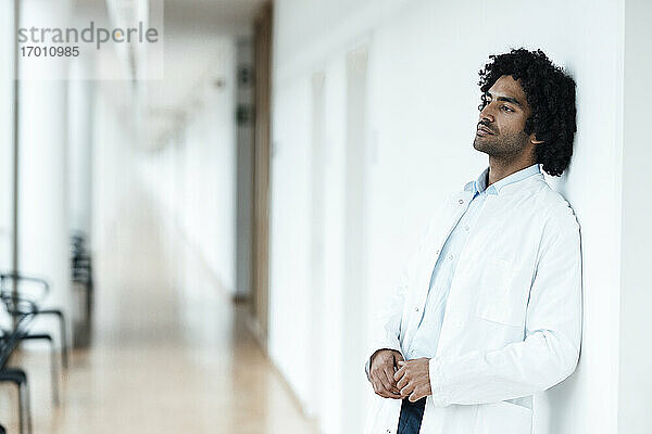 Nachdenklicher junger männlicher Arzt lehnt an einer weißen Wand im Krankenhausflur