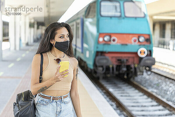 Junge Frau mit Gesichtsmaske  die wegschaut  während sie am Bahnhof steht und ein Mobiltelefon benutzt