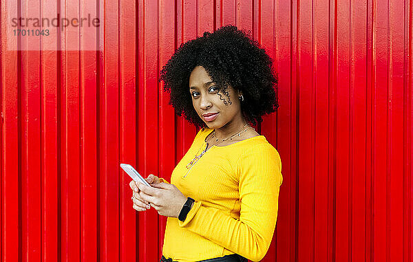 Selbstbewusste schöne Frau mit Afro-Haar  die ein Mobiltelefon vor einer roten Wand benutzt