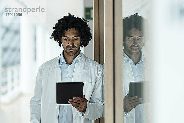 Junger männlicher Arzt  der ein digitales Tablet benutzt und sich an eine Glaswand im Krankenhaus lehnt