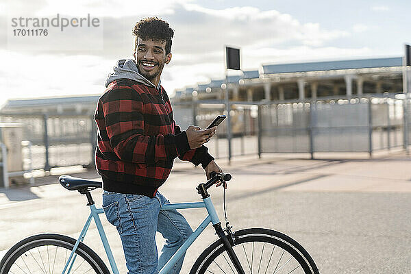 Lächelnder Mann mit Mobiltelefon  der wegschaut  während er an einem sonnigen Tag auf dem Fahrrad steht