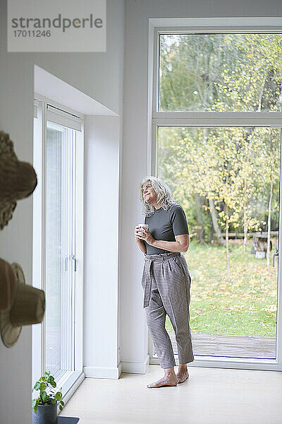 Nachdenkliche ältere Frau  die eine Tasse hält und durch das Fenster schaut  während sie sich zu Hause an die Wand lehnt