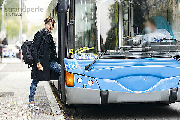 Lächelnder gut aussehender junger Mann mit Rucksack beim Einsteigen in einen Bus in der Stadt