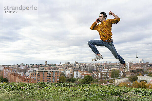 Aufgeregter junger Mann  der mit seinem Handy spricht  während er über einen Hügel vor einer Stadtlandschaft springt