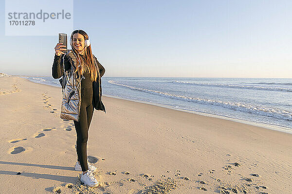 Lächelnde Frau nimmt Selfie mit Smartphone am Strand gegen klaren Himmel
