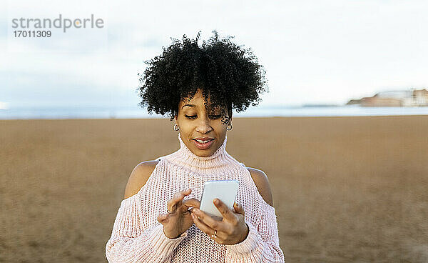 Frau mit Afro-Haar  die ein Mobiltelefon benutzt  während sie am Strand gegen den Himmel steht