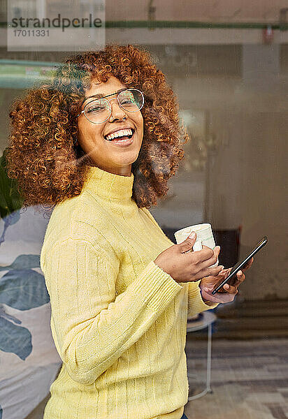 Lachende Frau mit Mobiltelefon beim Kaffee trinken hinter Glas in einem Cafe