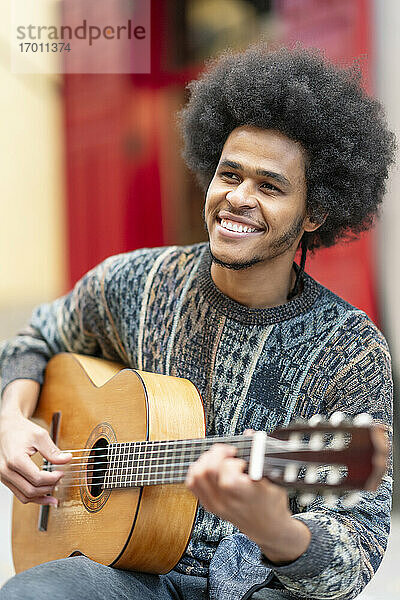 Lächelnder junger Mann mit lockigem Haar  der im Freien sitzend Gitarre spielt