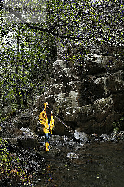 Frau in gelbem Regenmantel mit geschlossenen Augen steht am Bach im Wald