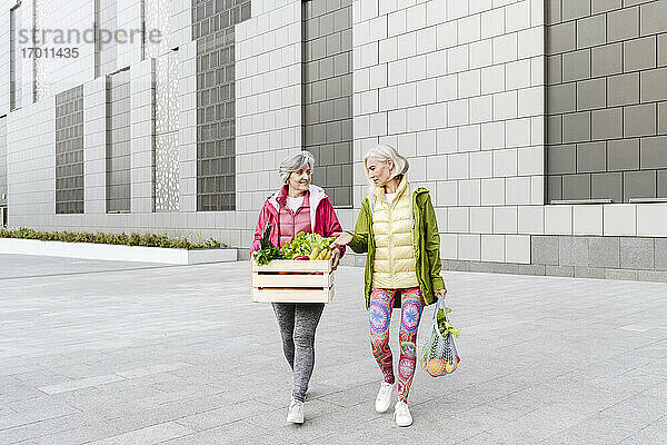 Freundinnen gehen mit Holzkiste und Netzbeutel mit Lebensmitteln auf dem Fußweg gegen die Wand