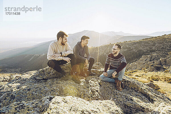 Männliche Freunde unterhalten sich auf einem Berg sitzend gegen einen klaren Himmel an einem sonnigen Tag