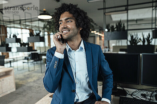 Lächelnder junger Geschäftsmann  der mit seinem Smartphone telefoniert  während er am Arbeitsplatz wegschaut