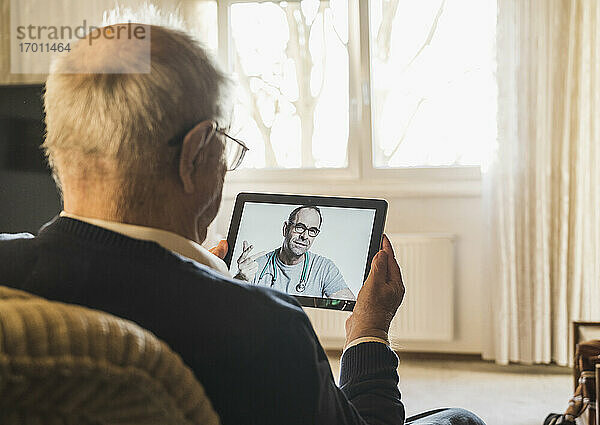 Männlicher Allgemeinmediziner berät einen älteren Mann per Videoanruf über ein digitales Tablet im Wohnzimmer