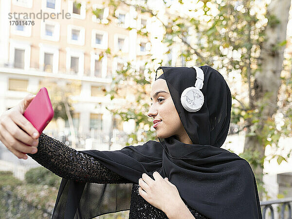 Porträt einer jungen schönen Frau mit Hidschab und Kopfhörern  die ein Selfie mit ihrem Smartphone macht