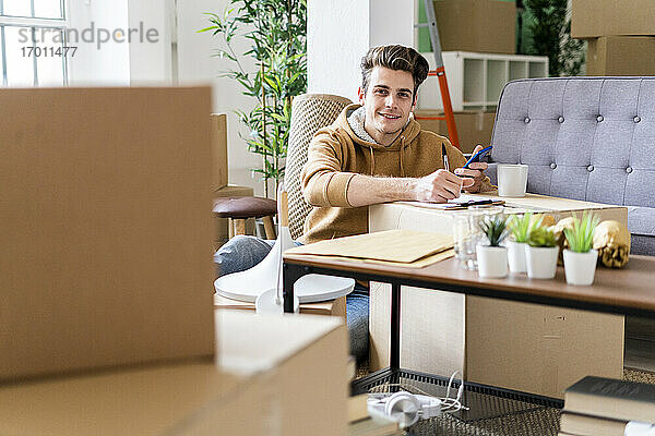 Lächelnder Mann mit Kugelschreiber  der ein Mobiltelefon über einer Box in einer neuen Wohnung hält
