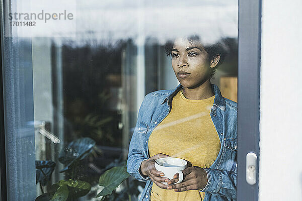 Frau schaut durch ein Fenster  hält eine Kaffeetasse
