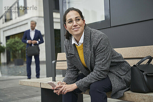 Lächelnde Geschäftsfrau auf einer Bank sitzend  die an einer Bushaltestelle wartet  während ein Geschäftsmann im Hintergrund steht