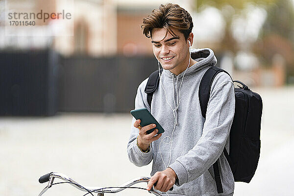 Lächelnder  modischer junger Mann  der sein Smartphone benutzt  während er in der Stadt Fahrrad fährt