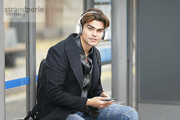 Hübscher junger Mann hört Musik  während er an der Bushaltestelle sitzt
