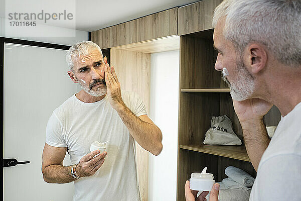 Älterer Mann trägt Gesichtscreme auf  während er im Badezimmer in den Spiegel schaut