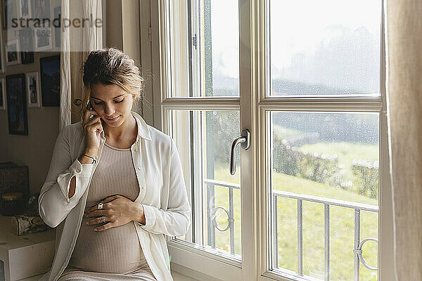 Junge schwangere Frau mit Hand auf dem Bauch  die zu Hause über ihr Smartphone spricht