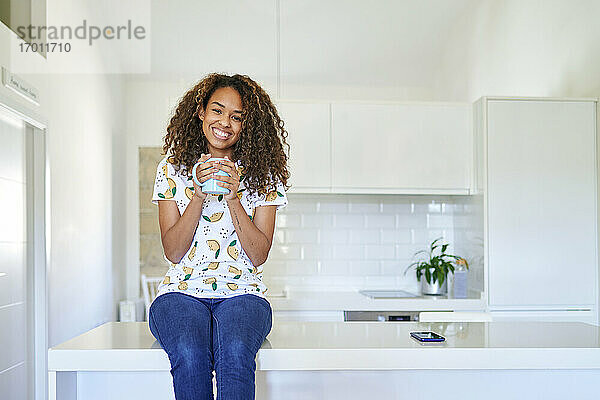 Lächelnde junge Frau mit Kaffeetasse auf dem Küchentisch zu Hause