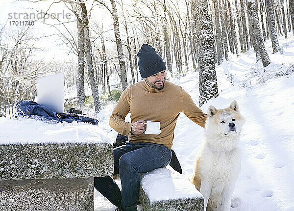 Lächelnder Mann hält Kaffeetasse und streichelt seinen Hund im verschneiten Wald