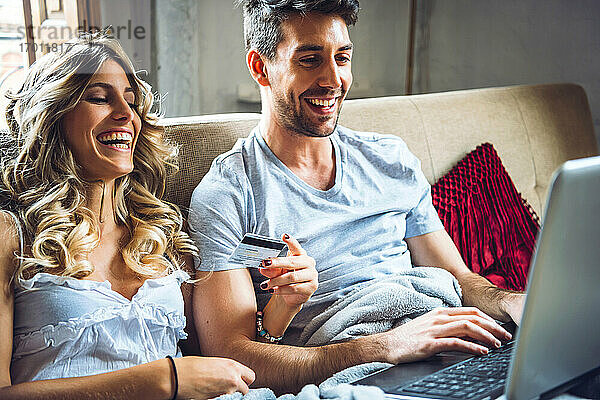 Glückliches junges Paar sitzt zu Hause auf der Couch mit Kreditkarte und Laptop