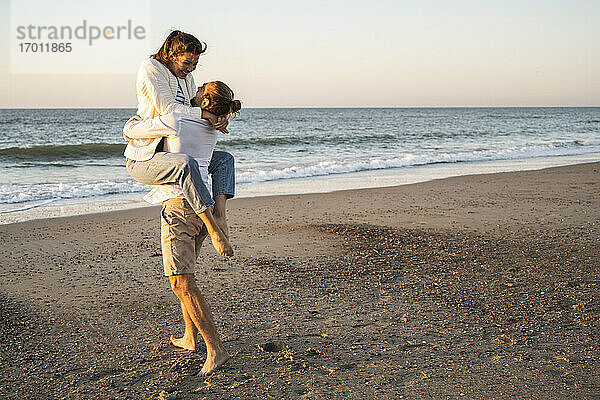 Unbeschwerter junger Mann hebt Freundin am Strand bei Sonnenuntergang