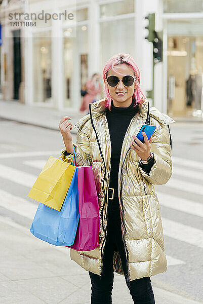 Lächelnde Frau  die Einkaufstüten trägt  während sie ein Mobiltelefon benutzt  in der Stadt stehend