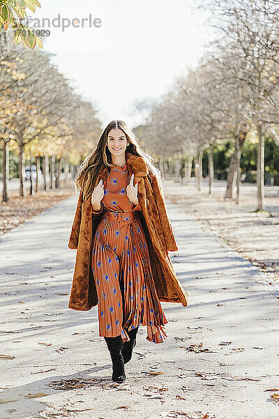 Lächelnde modische Frau mit Pelzmantel  die an einem sonnigen Tag auf einem Fußweg spazieren geht